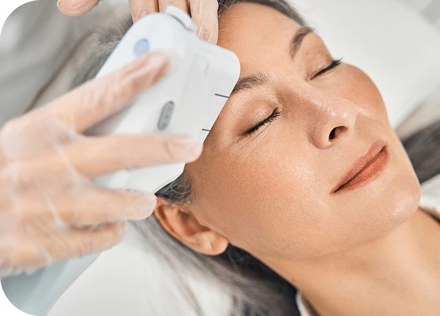 A woman receiving a facial treatment with a handheld device in a clinical setting, her eyes closed and relaxed.
