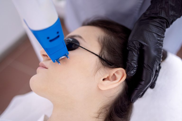 A close-up of a woman wearing glasses receiving a treatment with a blue device while being assisted by a professional.