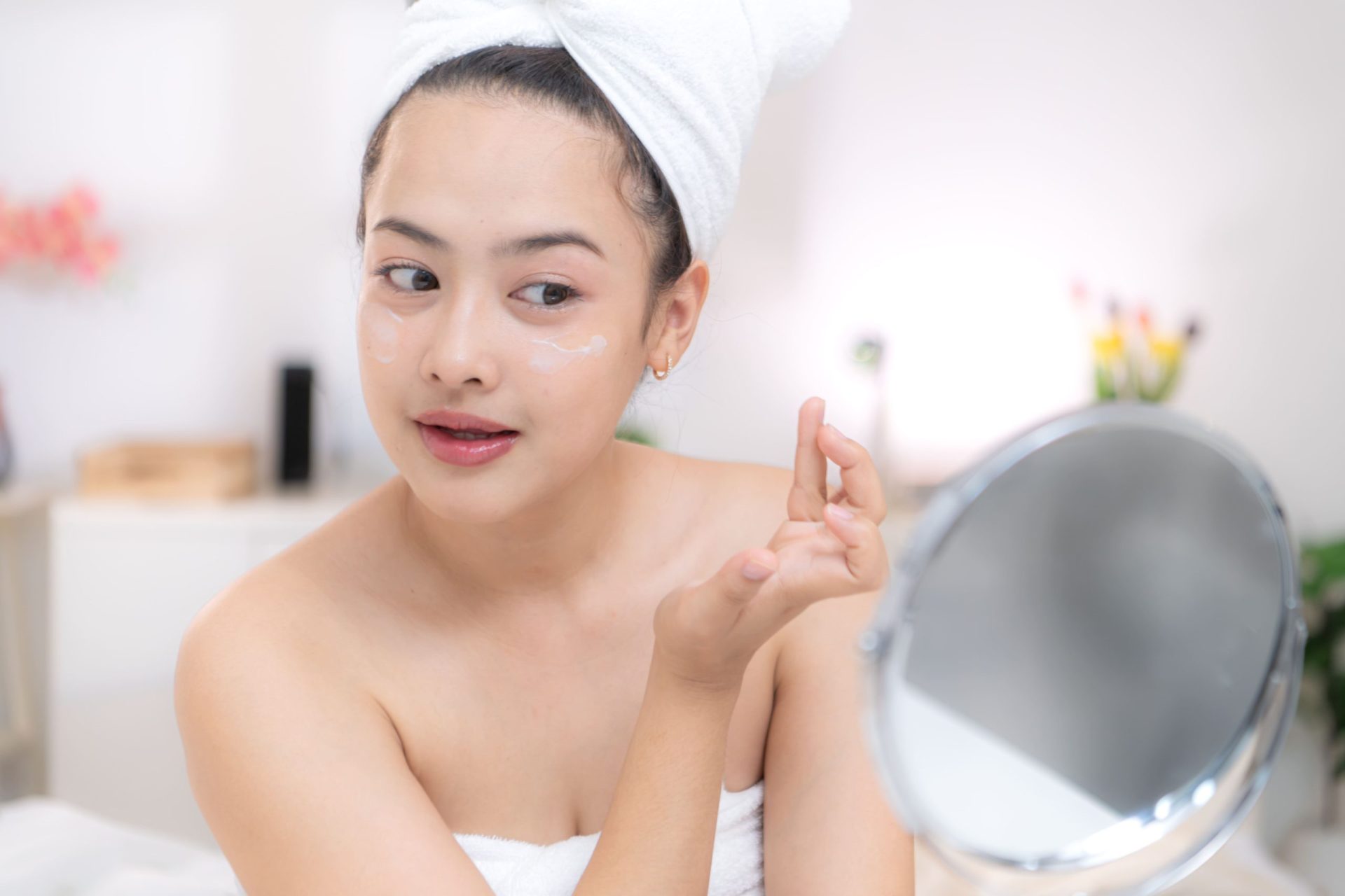 A young woman applying moisturizer to her face in a brightly lit bathroom.