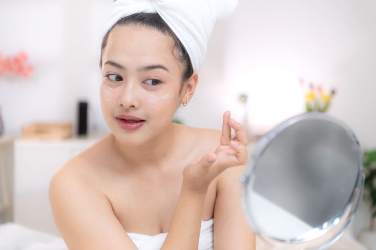 A young woman applying moisturizer to her face in a brightly lit bathroom.