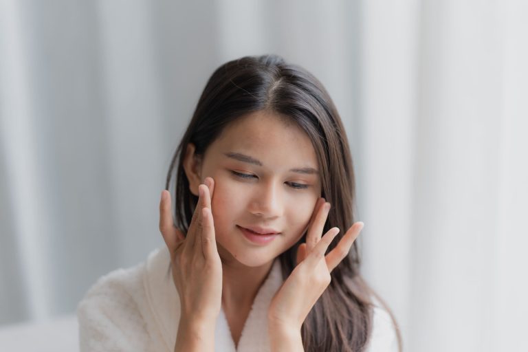 A young woman in a bathrobe applying skincare to her face in a bright and serene environment.